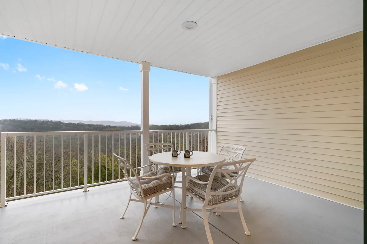 An inviting private balcony featuring an outdoor dining table set for two, with two coffee cups placed on the table. The balcony overlooks rolling green hills, lush trees, and distant mountain views. The sturdy railing frames the view, and the soft natural light suggests either an early morning or late afternoon setting.