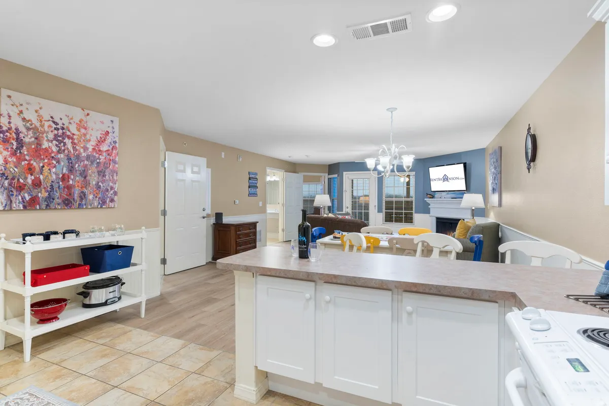 A cheerful, modern kitchen with a central island and colorful chairs stands adjacent to the living area. The kitchen is fully equipped, and vibrant artwork accents the walls. The fireplace and TV remain visible, visually connecting the kitchen space to the living and dining areas for a seamless, open-concept feel.