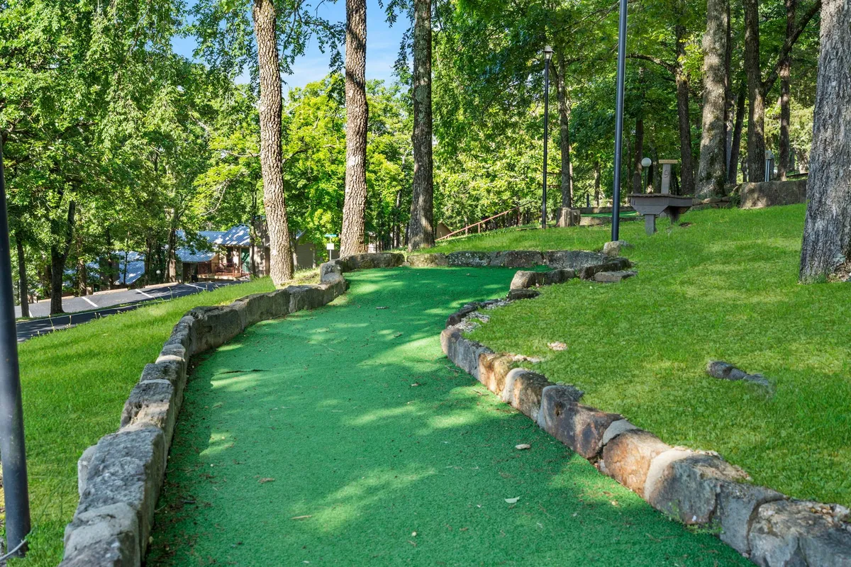 A playful mini-golf pathway winds through lush greenery and stone borders, surrounded by tall trees where families and friends are seen enjoying an engaging game. The setting is vibrant and lively, inviting group activities and friendly competition.
