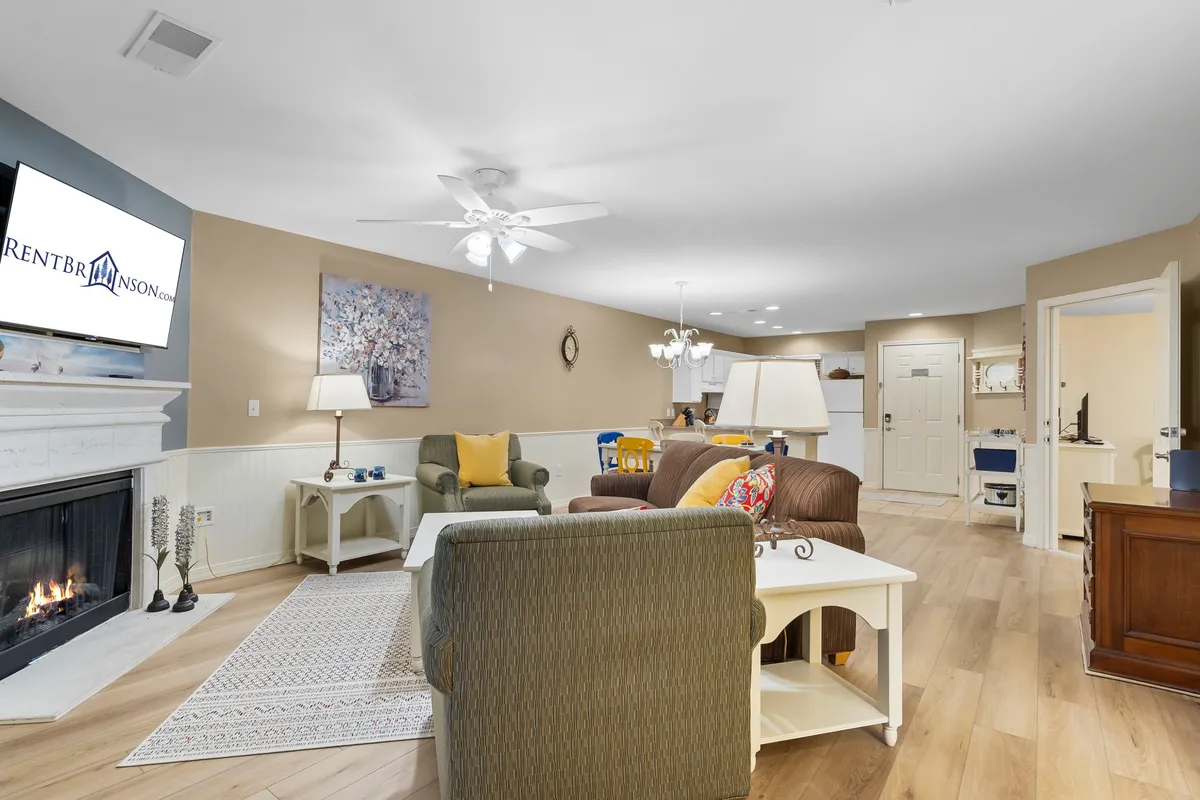 This image features a spacious, open-concept living and dining area with a cozy fireplace as the focal point. Comfortable seating is arranged for family gatherings, with a television above the fireplace, artwork on the walls, and a dining table set nearby. Sunlight fills the space, creating a warm and inviting atmosphere ideal for both relaxing and socializing.