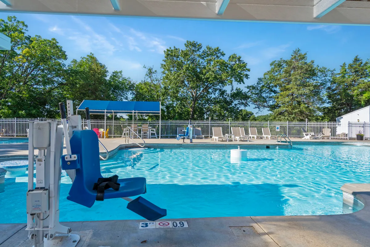 A sparkling outdoor swimming pool is bordered by lounge chairs and lush greenery, with a wheelchair lift visibly ensuring accessibility. The blue sky and surrounding trees create an inviting, relaxed atmosphere ideal for gathering and recreation.
