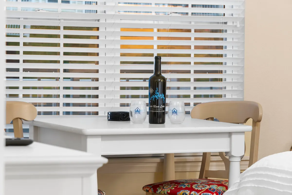 A close-up of a wine bottle and two glasses set on a white table, suggesting a cozy, intimate moment. Natural light hints at a picturesque view beyond the window, evoking a sense of winding down after an active day.