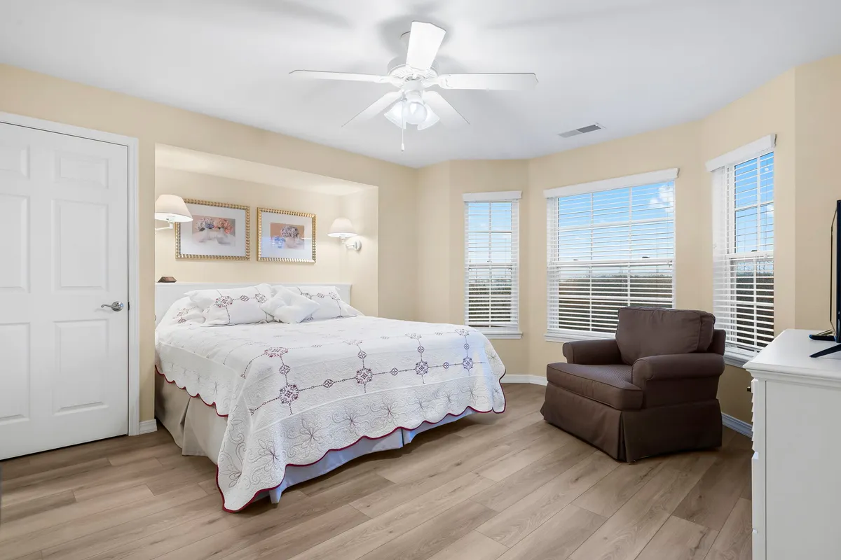 A tranquil bedroom featuring a king-sized bed adorned with a decorative quilt, a comfortable armchair situated by large windows that allow natural light to pour in, and a calm, neutral color palette. The room exudes an inviting and restful ambiance, ideal for unwinding after a busy day.