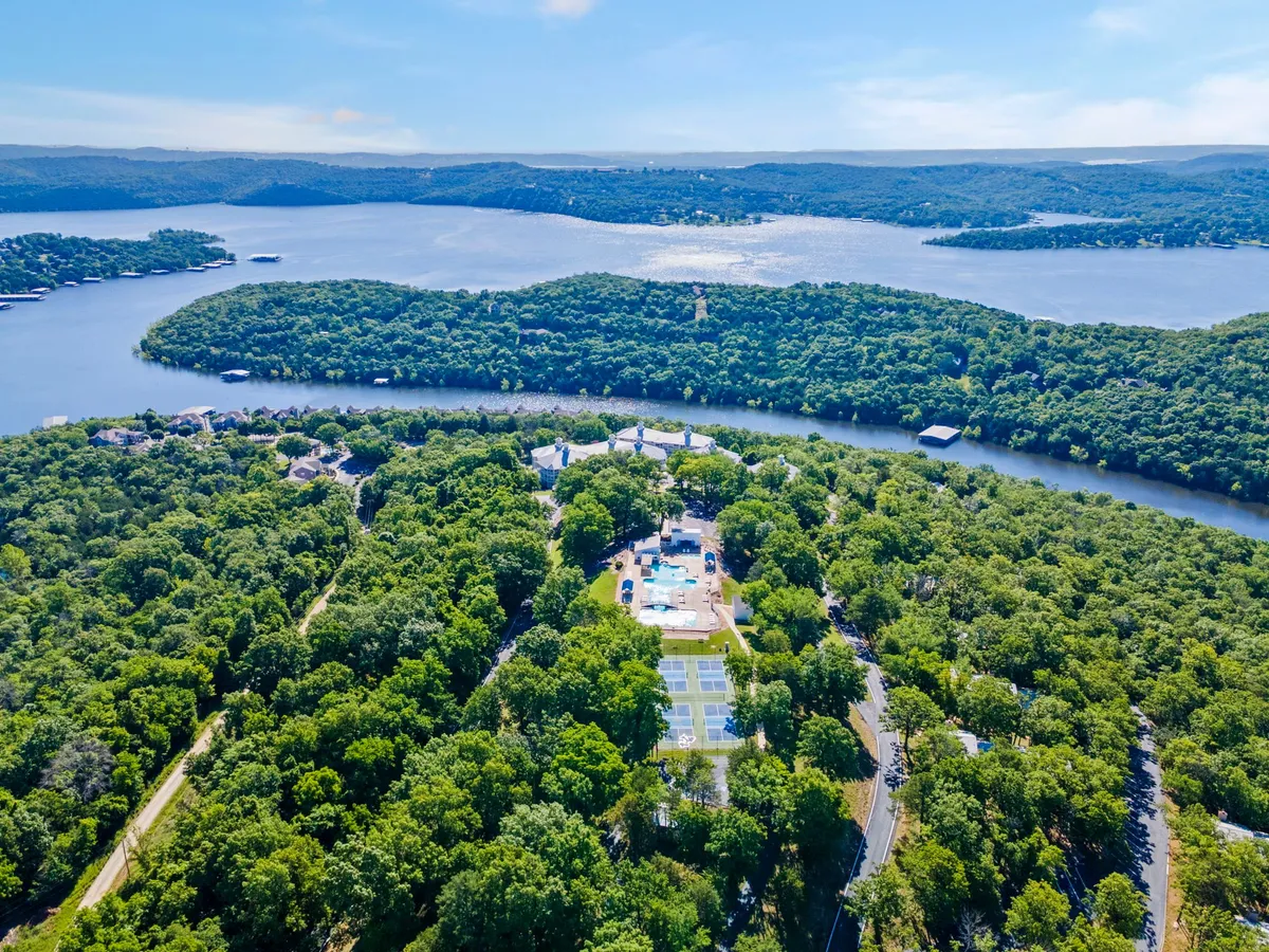 An expansive view reveals a sparkling swimming pool area, tennis courts, and lush greenery set against a stunning water backdrop. The scene captures a range of recreational amenities, including spaces for swimming, tennis, and lakeside relaxation, all nestled within a vibrant, active resort setting.