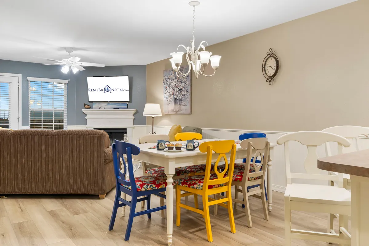 A central dining table surrounded by colorful chairs takes center stage, with the fireplace and TV visible in the background. The scene is bright and cheerful, set up for family meals, holiday gatherings, or memorable game nights, reinforcing the importance of shared experiences around the table.